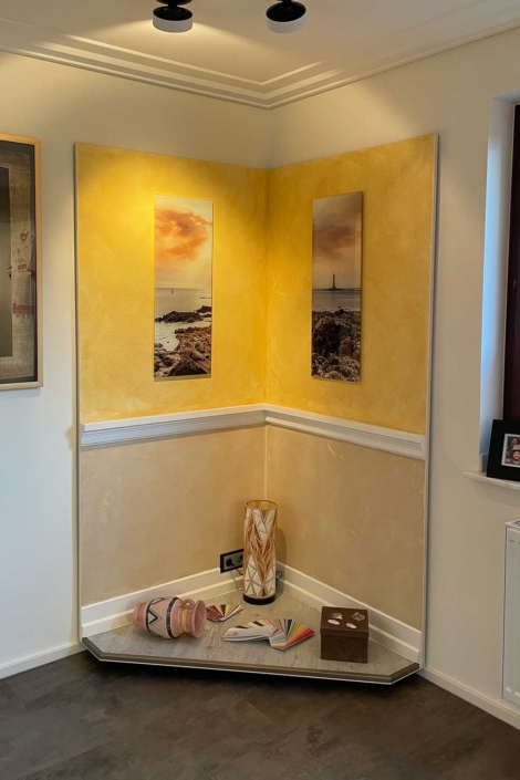Dekorative Wandecke im Showroom des Malerfachbetriebs Binder mit gestalteten Wandflächen in Gelb-Beige, Bildern, Zierleiste und Podest mit Dekorationselementen