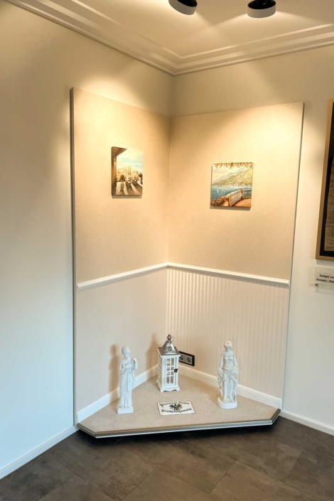 Dekorativ gestaltete Showroom-Koje des Malerfachbetriebs Binder mit cremefarbener Wandstruktur, Zierleiste, mediterranen Bildern und klassischen Dekoobjekten